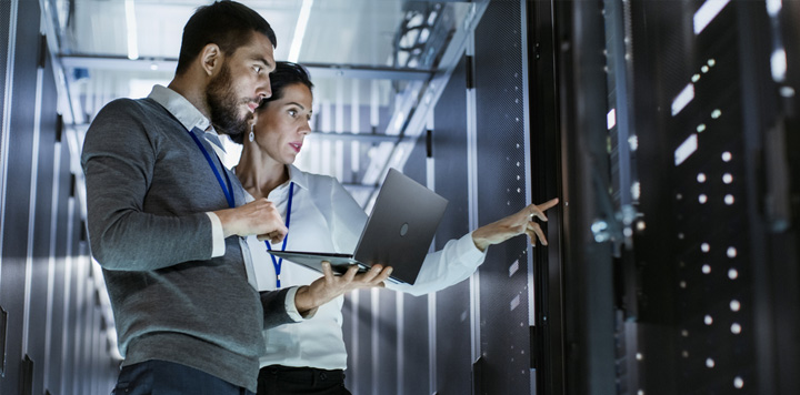 Two people engaged with a laptop in a server room, surrounded by rows of servers and technical equipment.