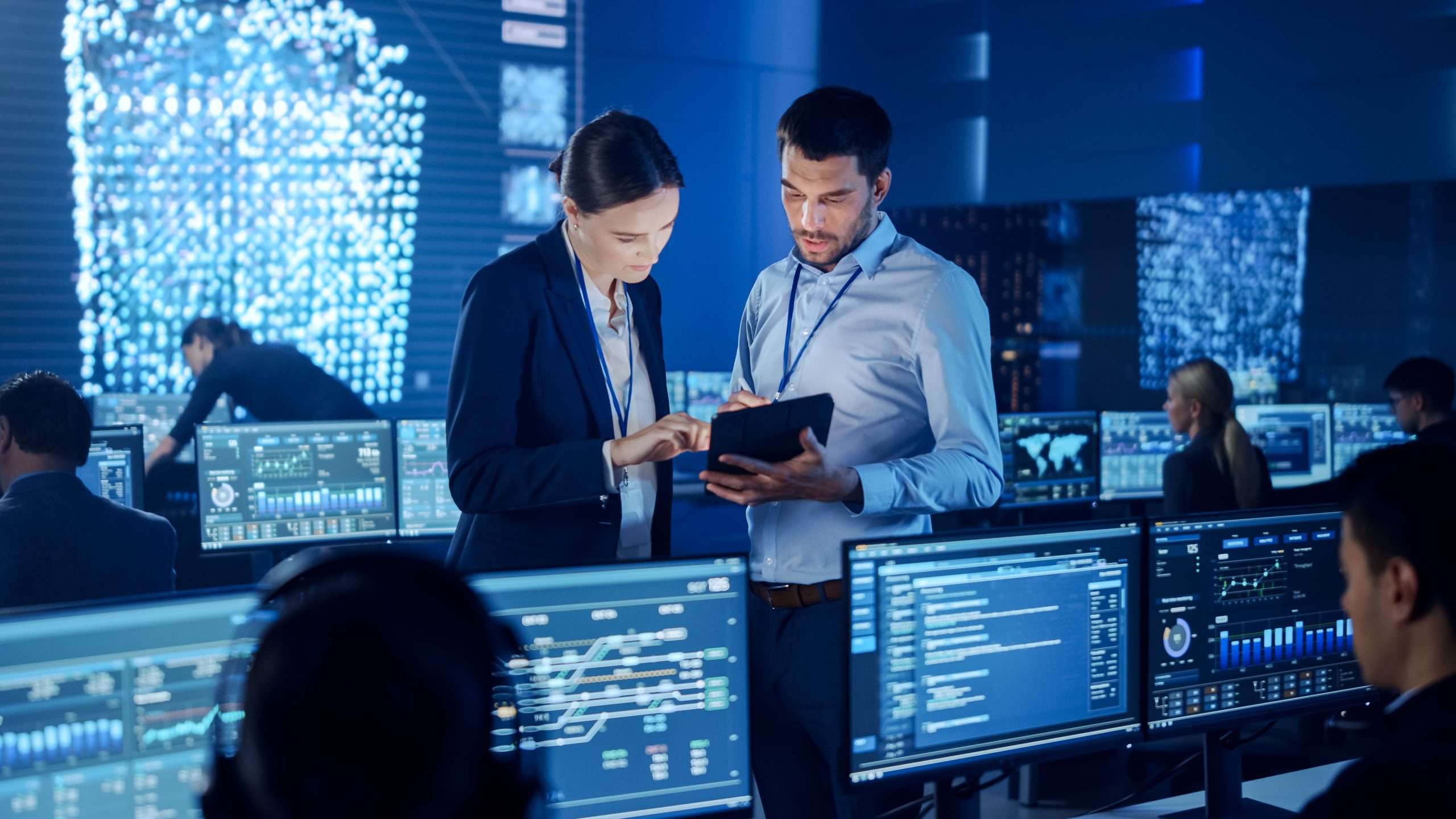 Two professionals in a tech-filled room, surrounded by multiple computers and monitors, concentrating on their work.